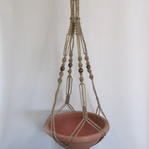 May include: A macrame plant hanger made of natural jute rope with brown wooden beads. The hanger is designed to hold a terracotta pot.