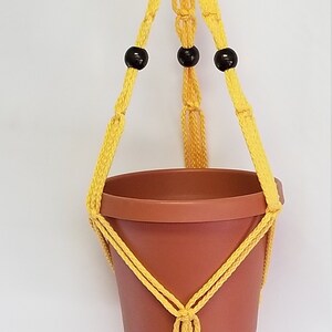 May include: A yellow macrame plant hanger with three black wooden beads. The hanger is holding a terracotta pot.