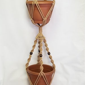 May include: A double-tiered macrame plant hanger with brown wooden beads. The hanger is made of natural jute rope and has a tassel at the bottom.