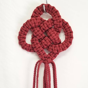 May include: A red macrame knot with a tassel hanging from the bottom. The knot is made with a thick cord and is tied in a complex pattern.
