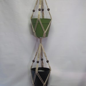 May include: A macrame plant hanger with two pots, one green and one black, hanging from a white ceiling. The hanger has wooden beads.