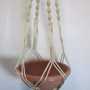 May include: A macrame plant hanger with a cream-colored rope and a brown terracotta pot. The hanger is made with a knotting technique and has a loop at the top for hanging.