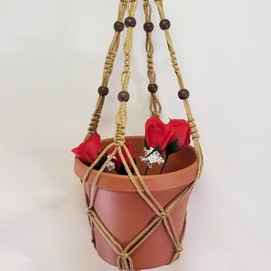 May include: A macrame plant hanger with brown wooden beads and a terracotta pot with red flowers. The hanger has a tassel at the bottom.