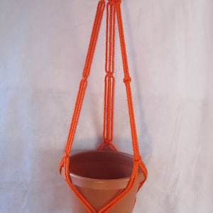 May include: Orange macrame plant hanger with a terracotta pot. The hanger is made of thick cord and has a tassel at the bottom.