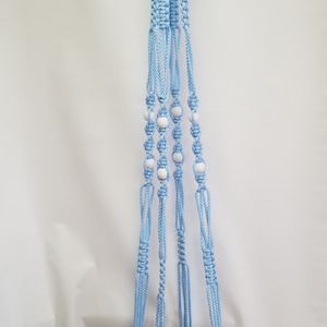 May include: A light blue macrame plant hanger with white beads. The plant hanger holds a terracotta pot filled with red and white flowers. The hanger is suspended from a gold ring.