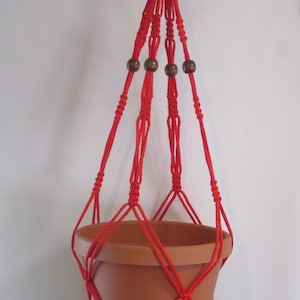 May include: Red macrame plant hanger with wooden beads. The hanger is holding a terracotta pot.