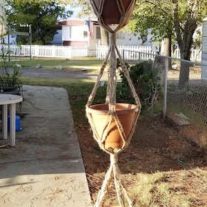 May include: A three-tiered macrame plant hanger with three terracotta pots. The hanger is made of natural jute rope and is hanging from a tree branch.