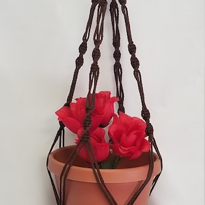 May include: A brown macrame plant hanger with a terracotta pot and red flowers. The hanger is made of cotton cord and has a knotted design.