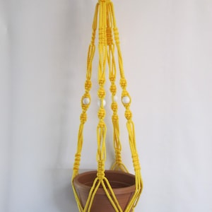May include: Yellow macrame plant hanger with white beads. The hanger is made of cotton cord and has a woven design. It is suitable for hanging a small plant pot.