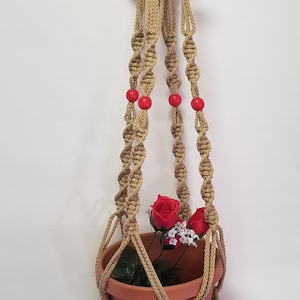 May include: A macrame plant hanger with a terracotta pot and artificial flowers. The hanger is made of natural jute rope and has red wooden beads.
