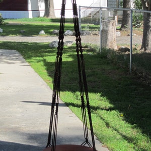 May include: A brown macrame plant hanger with a terracotta pot hanging from it. The hanger is suspended in front of a white mobile home with a red roof.