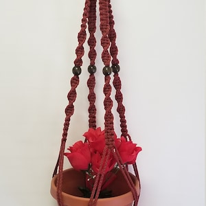 May include: A burgundy macrame plant hanger with wooden beads and a terracotta pot with red silk flowers.