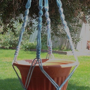 May include: A light blue macrame plant hanger with a terracotta pot. The hanger is made of knotted rope and has a tassel at the bottom.