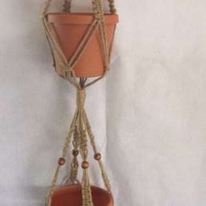 May include: A macrame plant hanger with two terracotta pots and wooden beads. The hanger is made of natural jute rope and has a tassel at the bottom.