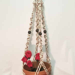May include: A white and brown macrame plant hanger with wooden beads. The hanger is holding a terracotta pot with red silk flowers.