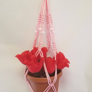 May include: A pink macrame plant hanger with white beads, holding a terracotta pot with red artificial flowers.
