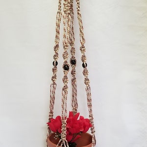 May include: A macrame plant hanger with a terracotta pot and red flowers. The hanger is made with white and brown rope and has black beads.