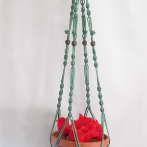 May include: A hanging macrame plant holder in a soft green color, with a terracotta-colored pot filled with red flowers. The macrame design features wooden beads and a tassel at the bottom. The plant holder is suspended from a gold-colored ring.