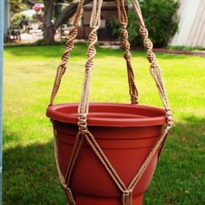 May include: A brown macrame plant hanger with a terracotta pot hanging from it. The hanger is made of thick rope and has a tassel at the bottom.