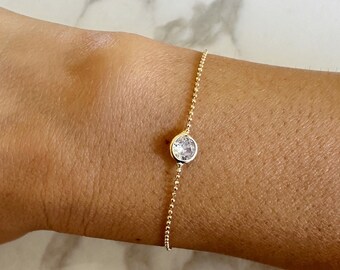 May include: A delicate gold bracelet featuring a single, round, clear gemstone. The bracelet's chain is composed of small, evenly spaced beads. The gemstone is set in a simple bezel setting, creating a minimalist and elegant design.