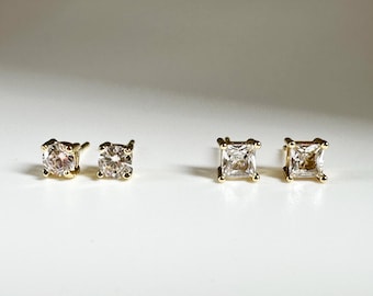 May include: Two pairs of gold stud earrings. One pair features round-cut clear gemstones, and the other pair has square-cut clear gemstones. The earrings are set in gold prongs, creating a classic and elegant look.