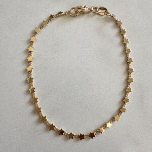 May include: A gold-toned chain bracelet featuring a continuous series of small, flat, five-pointed star charms linked together. The bracelet is secured with a small lobster claw clasp at the top, creating a circular shape with a polished finish.