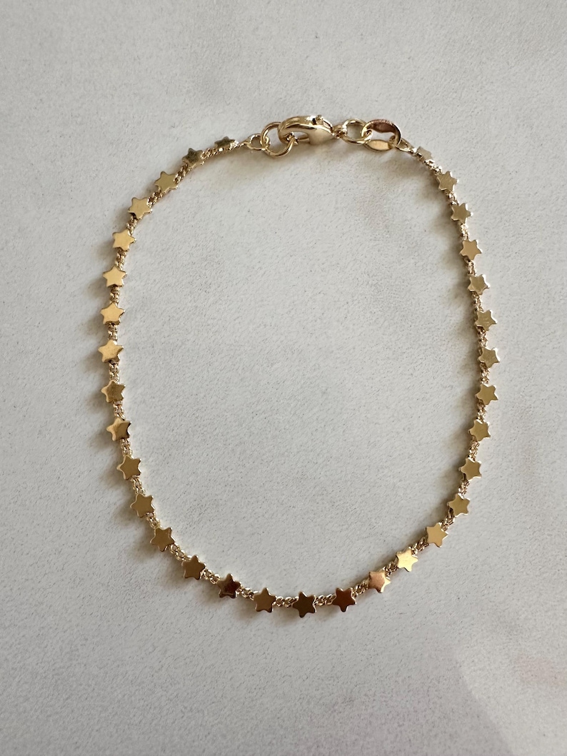 May include: A gold-toned chain bracelet featuring a continuous series of small, flat, five-pointed star charms linked together. The bracelet is secured with a small lobster claw clasp at the top, creating a circular shape with a polished finish.