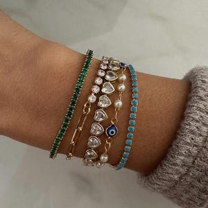 May include: A collection of gold bracelets adorns a wrist. The bracelets include a green gemstone tennis bracelet, a gold chain, a bracelet with heart-shaped clear stones, a pearl and gold chain, and a turquoise bracelet with small stones.