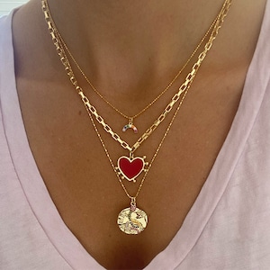 Może przedstawiać: Zestaw złotych naszyjników warstwowych. Zawiera zawieszkę w kształcie tęczy, czerwoną zawieszkę w kształcie serca i okrągłą zawieszkę z motywem jednorożca. Naszyjniki są noszone przez osobę w różowej koszulce.