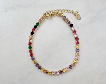 May include: A gold-toned bracelet with a row of colorful, round gemstones. The gemstones are arranged in a rainbow of colors, including red, orange, yellow, green, blue, and purple. The bracelet has a small heart-shaped charm.