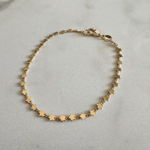 May include: A gold-toned chain bracelet featuring a series of small, evenly spaced star-shaped charms along the entire length of the chain. The bracelet includes a small lobster claw clasp mechanism for securing the piece around a wrist.