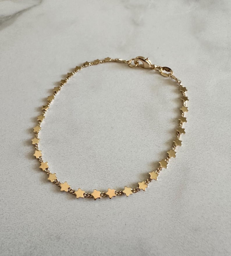 May include: A gold-toned chain bracelet featuring a series of small, evenly spaced star-shaped charms along the entire length of the chain. The bracelet includes a small lobster claw clasp mechanism for securing the piece around a wrist.