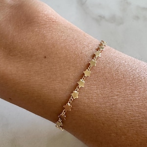 May include: A gold-toned chain bracelet featuring a series of small, uniform star-shaped charms linked together in a continuous row. The bracelet is designed with a delicate metal link construction that allows the star charms to lay flat.