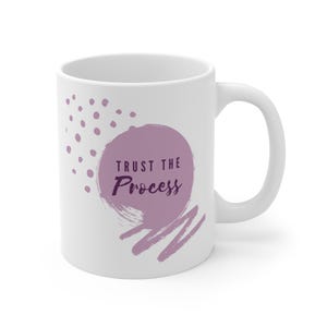 May include: White ceramic mug with a purple design that says "TRUST THE PROCESS". The design includes a purple circle, dots, and brushstrokes. The mug has a curved handle.