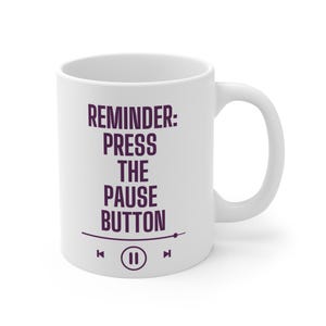 May include: White ceramic mug with the text "REMINDER: PRESS THE PAUSE BUTTON" in dark purple. The design includes a pause button symbol and music player controls. The mug has a curved handle.