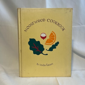 May include: A hardback cookbook titled Moosewood Cookbook by Mollie Katzen with a pale yellow cover. The front features a graphic illustration of a dark green leafy vegetable, a red radish, and an orange slice centred on the book cover.
