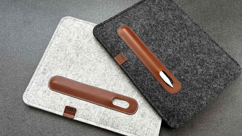 May include: Two felt iPad cases with a brown leather pen holder. The case on the left is light gray and the case on the right is dark gray.