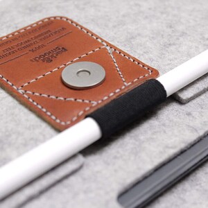 K&ouml;nnte beinhalten: Ein graues Filz-Etui mit einer braunen Lederklappe und einem schwarzen Gummiband. Die Klappe hat einen Metall-Druckknopfverschluss und der Text "UPSCALE" und "MODERN" ist darauf gedruckt. Ein wei&szlig;er Stift ist in das Gummiband gelegt.