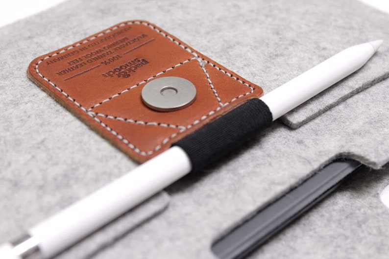 K&ouml;nnte beinhalten: Ein graues Filz-Etui mit einer braunen Lederklappe und einem schwarzen Gummiband. Die Klappe hat einen Metall-Druckknopfverschluss und der Text "UPSCALE" und "MODERN" ist darauf gedruckt. Ein wei&szlig;er Stift ist in das Gummiband gelegt.