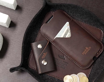 May include: A brown leather phone case with a card slot and the text "pack & smooch" embossed on the front. The case is sitting in a black felt tray with a key holder and some coins.
