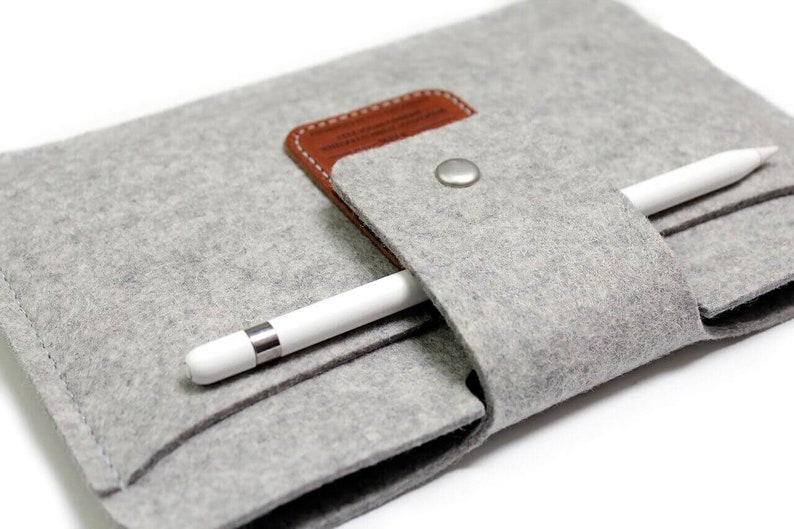 K&ouml;nnte beinhalten: Ein graues Filz-Etui mit einer braunen Lederklappe und einem Druckknopfverschluss. Ein wei&szlig;er Stift ist im Etui verstaut.