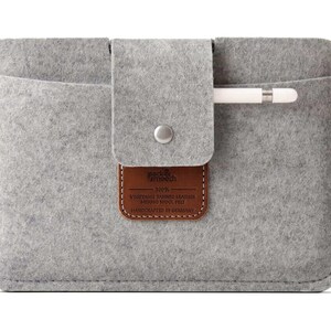 K&ouml;nnte beinhalten: Graue Filz-iPad-H&uuml;lle mit einem braunen Leder-Patch, auf dem "pad & pench 100% VEGETABLE TANNED LEATHER MERINO WOOL FELT HANDCRAFTED IN GERMANY" steht. Die H&uuml;lle hat ein Fach f&uuml;r einen Stift.