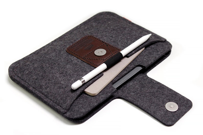 K&ouml;nnte beinhalten: Eine graue Filztasche mit einer braunen Lederklappe und einem Magnetverschluss. Ein wei&szlig;er Stift und ein Notizbuch befinden sich in der Tasche.