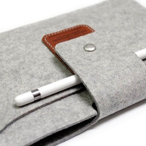 K&ouml;nnte beinhalten: Ein graues Filz-Etui mit einer braunen Lederklappe und einem silbernen Knopfverschluss. Ein wei&szlig;er Stift ist im Etui verstaut.