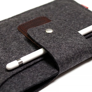 K&ouml;nnte beinhalten: Ein graues Filz-Etui mit einer braunen Lederklappe und einem silbernen Knopfverschluss. Ein wei&szlig;er Stift ist im Etui verstaut.
