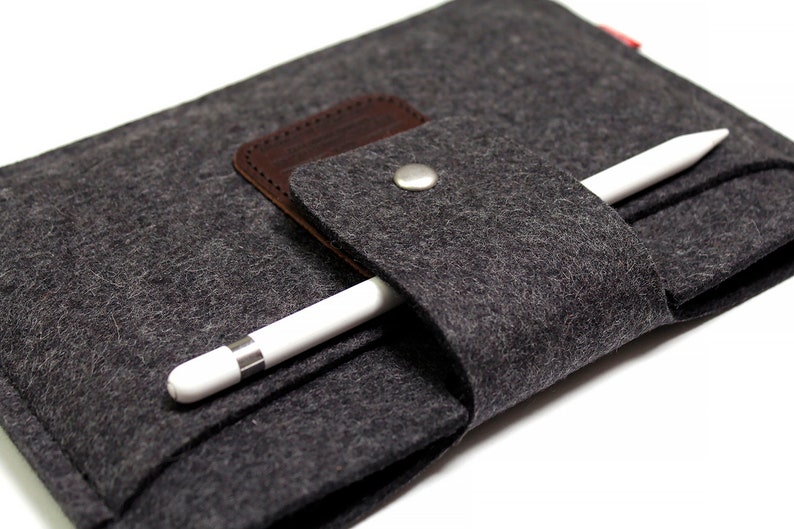 K&ouml;nnte beinhalten: Ein graues Filz-Etui mit einer braunen Lederklappe und einem silbernen Knopfverschluss. Ein wei&szlig;er Stift ist im Etui verstaut.