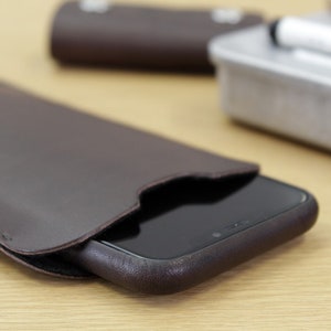 Op de afbeelding: Een bruine leren telefoonhoes met een zwarte telefoon erin. De hoes heeft een klep die het scherm van de telefoon bedekt.