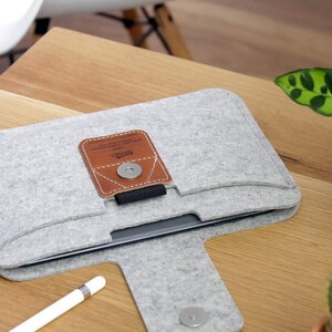 K&ouml;nnte beinhalten: Eine graue Filz-Laptoptasche mit einer braunen Lederklappe und einem Magnetverschluss. Die Tasche hat ein Fach auf der Vorderseite f&uuml;r einen Stift. Der Text "Field Notes" ist auf der Lederklappe aufgedruckt.