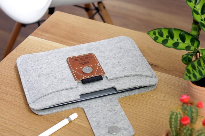 K&ouml;nnte beinhalten: Eine graue Filz-Laptoptasche mit einer braunen Lederklappe und einem Magnetverschluss. Die Tasche hat ein Fach auf der Vorderseite f&uuml;r einen Stift. Der Text "Field Notes" ist auf der Lederklappe aufgedruckt.