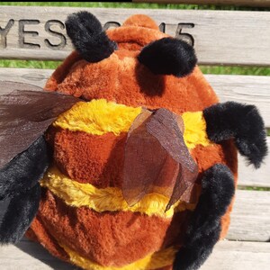 Könnte beinhalten: Ein Plüsch-Bienen-Spielzeug mit braunen und gelben horizontalen Streifen, schwarzen flauschigen Fühlern und durchscheinenden braunen Flügeln. Der Text "Brown Bee" ist in der oberen linken Ecke sichtbar. Das Spielzeug hat eine weiche Oberfläche.